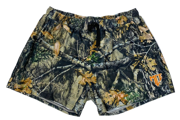 A pair of camouflage swim shorts featuring a woodland tree bark and leaf pattern in shades of brown, green, and black, with a small orange and white logo on the lower right leg, laid flat on a light gray background.